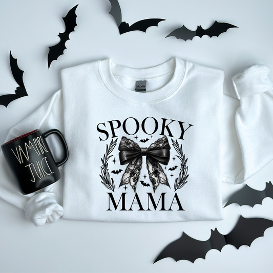 A white crewneck sweatshirt with 'Spooky Mama' text and a design featuring a black bow, bats, and a wreath, presented on a surface with bat cut-outs and a mug with 'BAT JUICE' text.