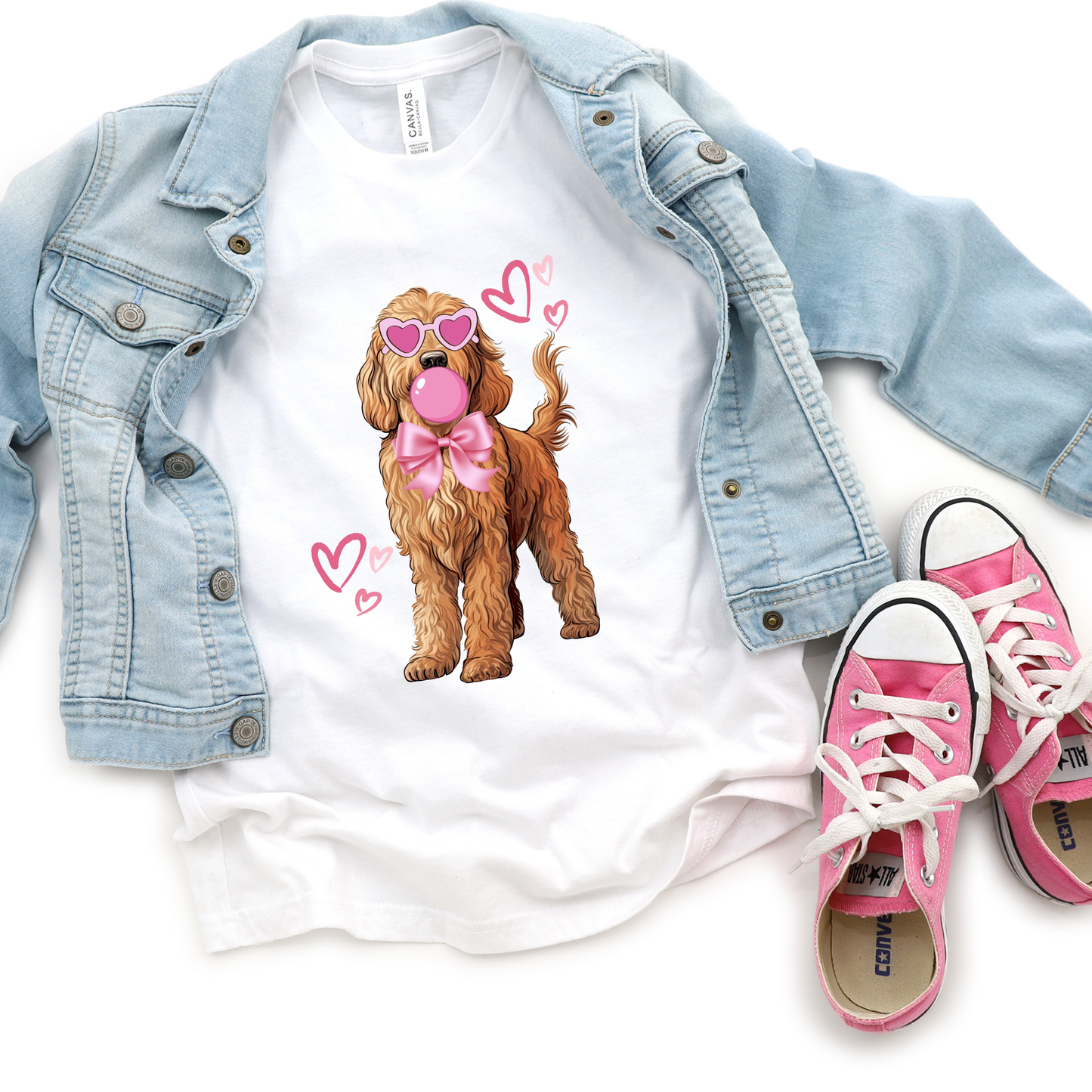 A white crew neck sweatshirt with a printed design of a cute, cartoon-style dog wearing pink sunglasses and a bow, surrounded by pink hearts. The sweatshirt is presented with a pair of pink and white sneakers and a blue denim jacket.