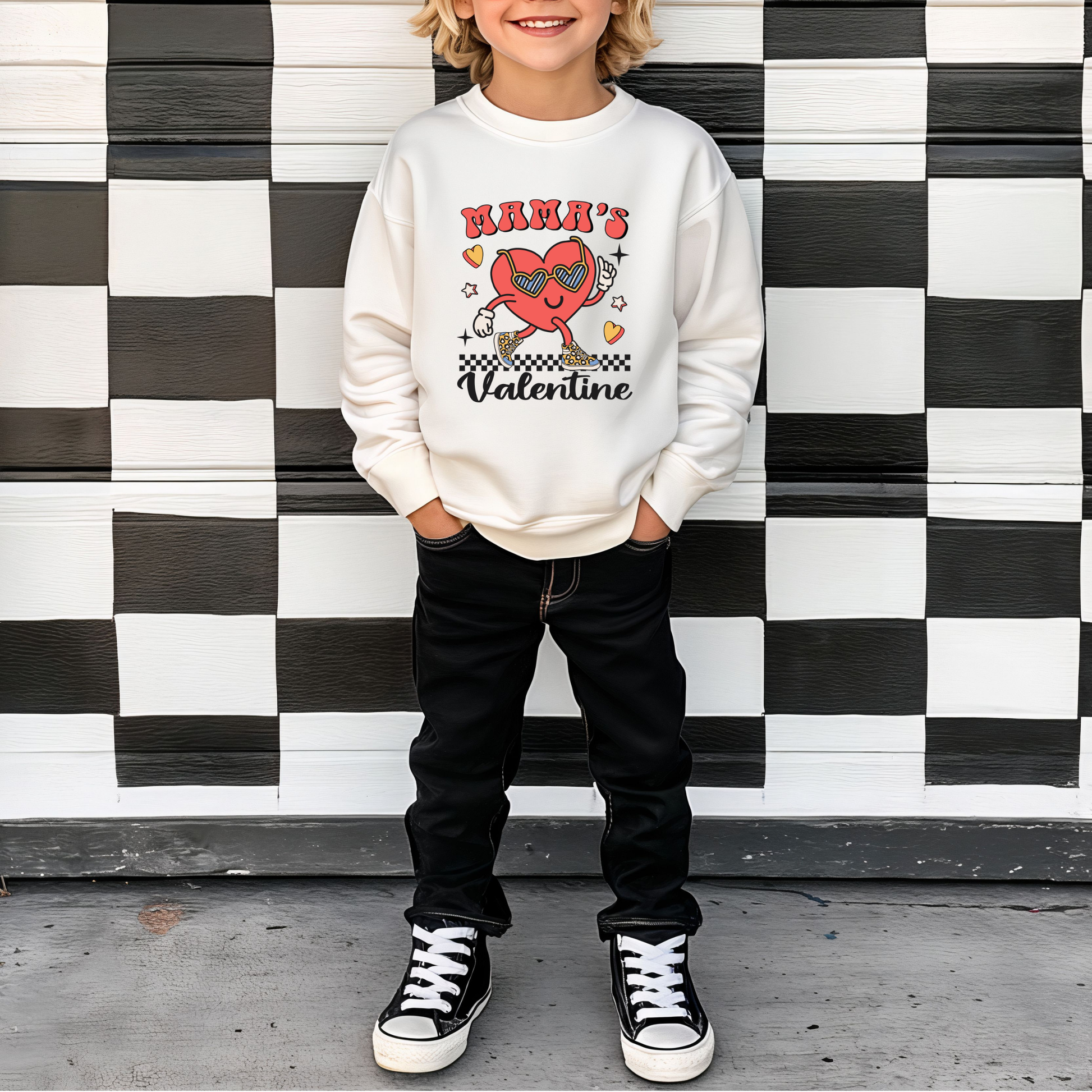 A child standing in front of a checkered background wearing a white sweatshirt with 'Mama's Valentine' text and a heart graphic character on it.