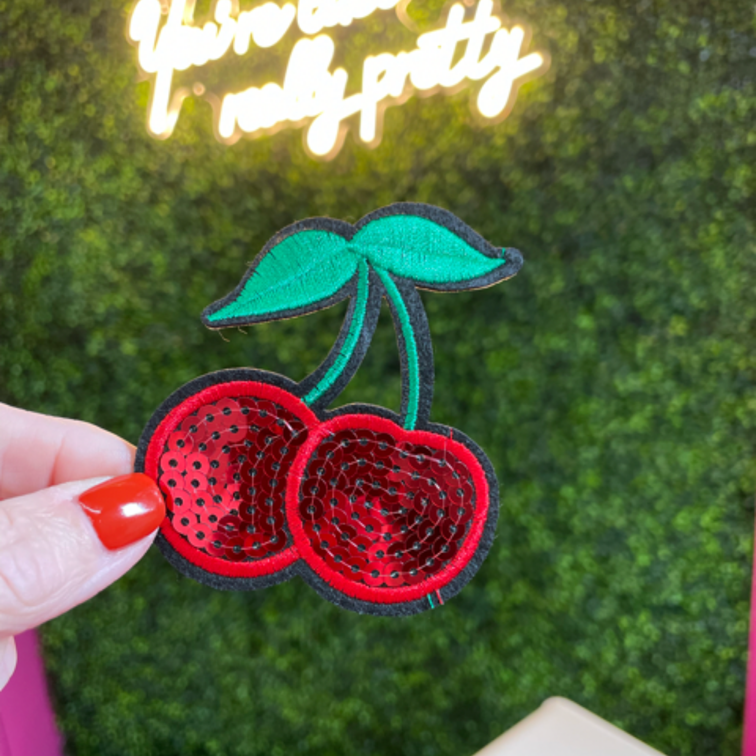 hand holding a Sequins cherry patch with a leafy backdrop and a neon sign