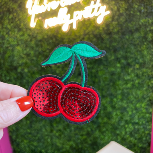 hand holding a Sequins cherry patch with a leafy backdrop and a neon sign