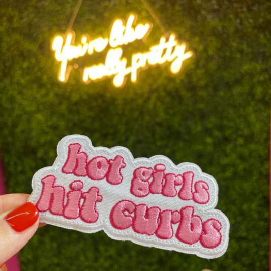 hand holding a pink embroidered patch that says hot girls hit curbs with a green leafy background. 
