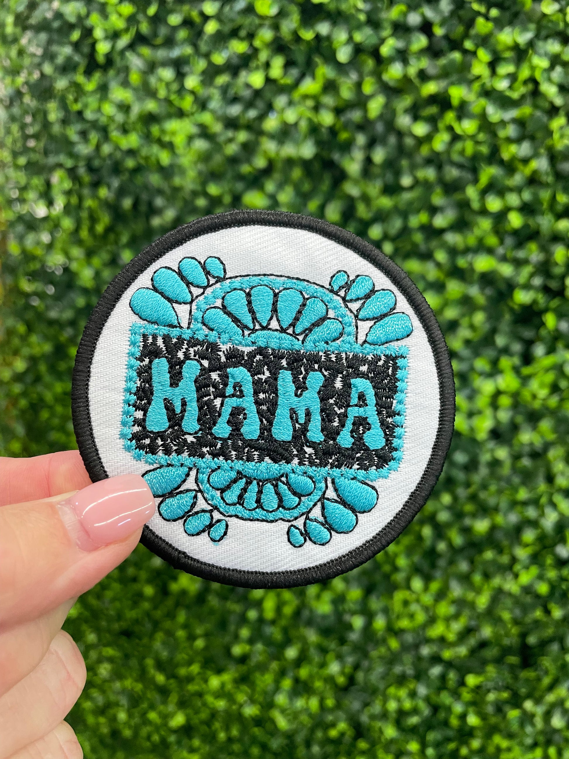 Circular patch with 'MAMA' text and teal designs held against a green textured background