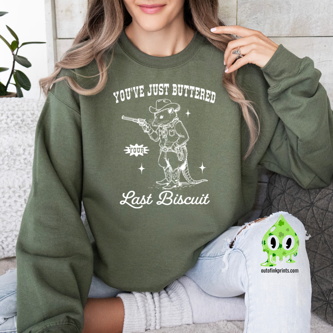 A person wearing a dark green sweatshirt with the text 'You've Just Buttered Last Biscuit' and an illustration of a mouse holding a gun.
