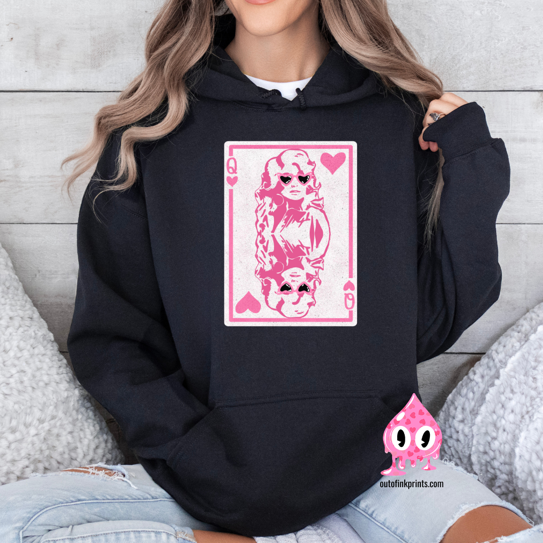 A woman sitting wearing a black hoodie with a pink graphic of Dolly Parton and the text 'Queen of Hearts' on it.