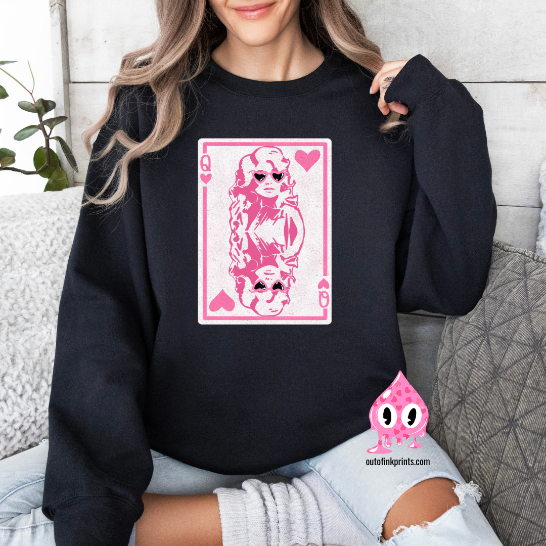 A person wearing a black sweatshirt with a pink graphic print of Dolly Parton and the text 'Queen of Hearts'. The sweatshirt is displayed in a cozy, casual setting with the brand's logo visible.