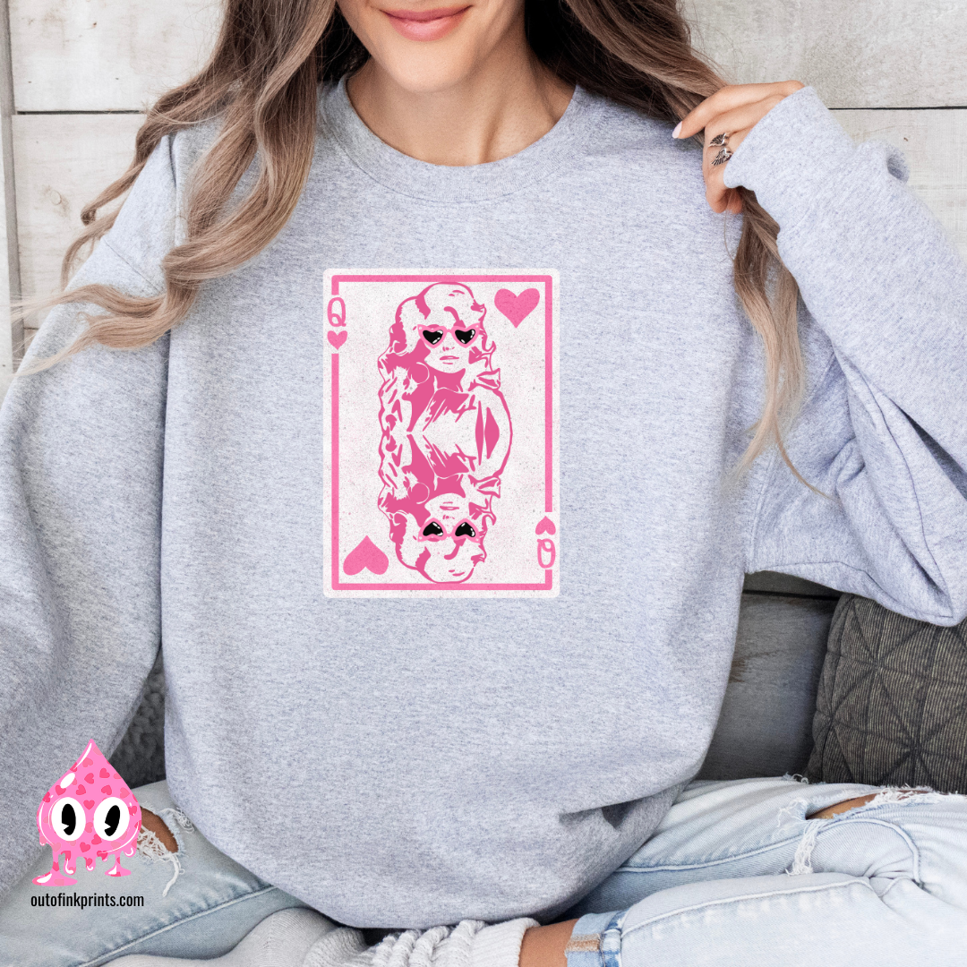 A person wearing a gray sweatshirt with a pink graphic print of Dolly Parton and the text 'Queen of Hearts'. The sweatshirt is displayed in a cozy, casual setting with the brand's logo visible.