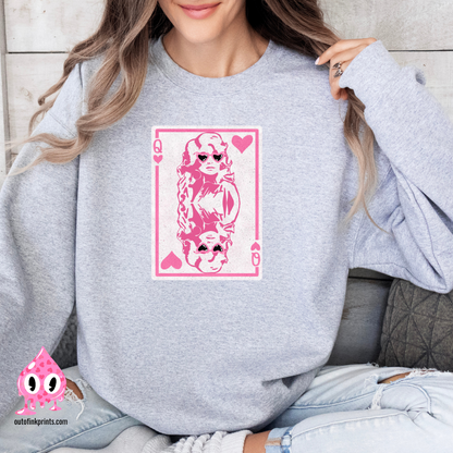 A person wearing a gray sweatshirt with a pink graphic print of Dolly Parton and the text 'Queen of Hearts'. The sweatshirt is displayed in a cozy, casual setting with the brand's logo visible.
