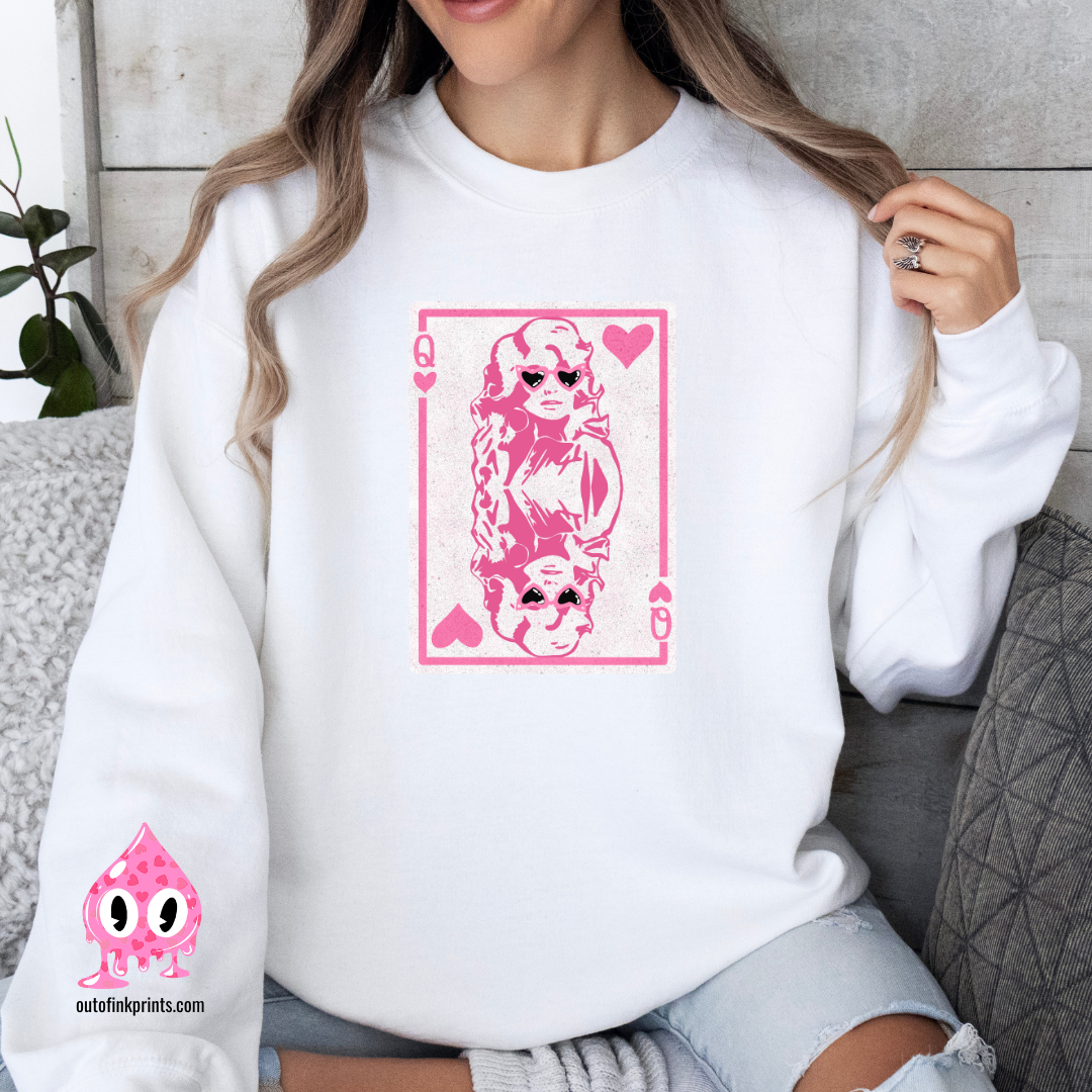 A person wearing a white sweatshirt with a pink graphic print of Dolly Parton and the text 'Queen of Hearts'. The sweatshirt is displayed in a cozy, casual setting with the brand's logo visible.