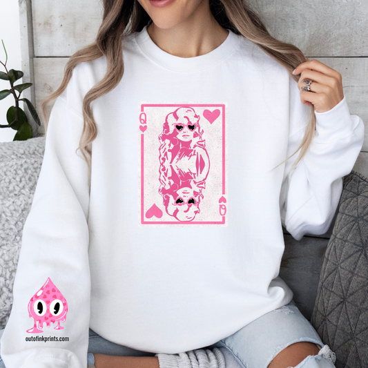A person wearing a white sweatshirt with a pink graphic print of Dolly Parton and the text 'Queen of Hearts'. The sweatshirt is displayed in a cozy, casual setting with the brand's logo visible.