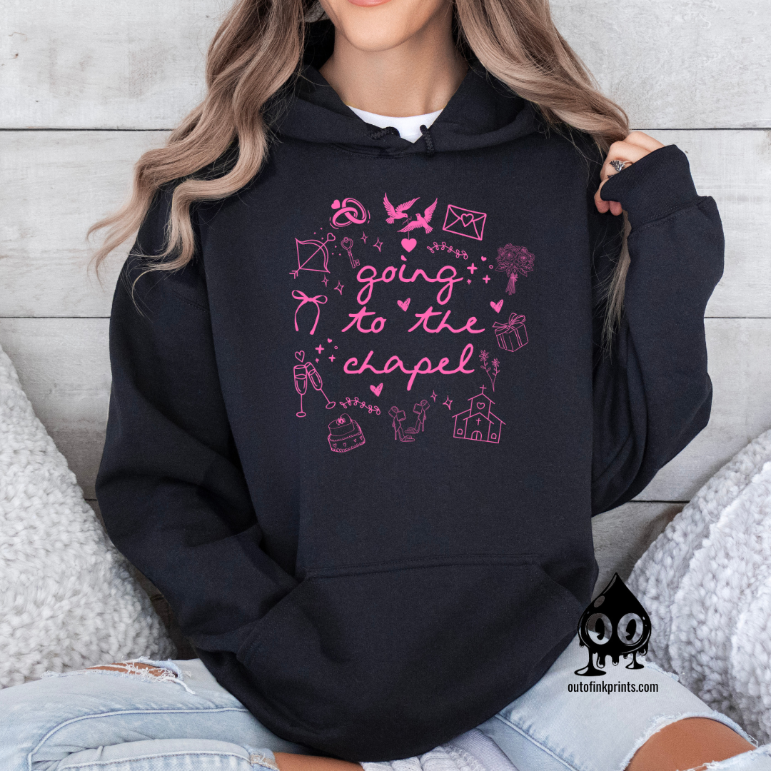 A woman wearing a black hoodie with pink text and graphics including bows, a bird, and a cake, sitting on a cozy white surface with wooden background.
