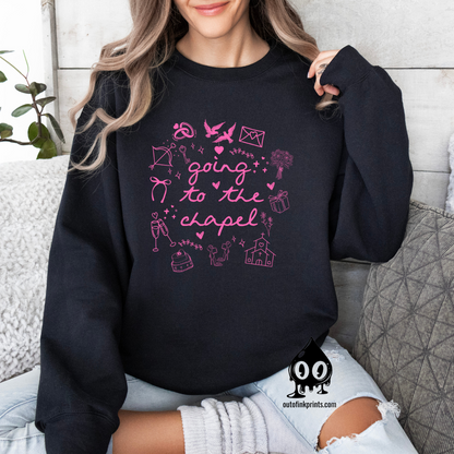 A woman sitting and wearing a black sweatshirt with pink text that reads 'Going to the Chapel' along with various symbols and text related to weddings.