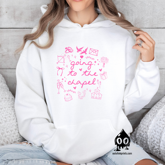 A woman wearing a white hoodie with pink text and graphics including bows, a bird, and a cake, sitting on a cozy white surface with wooden background.