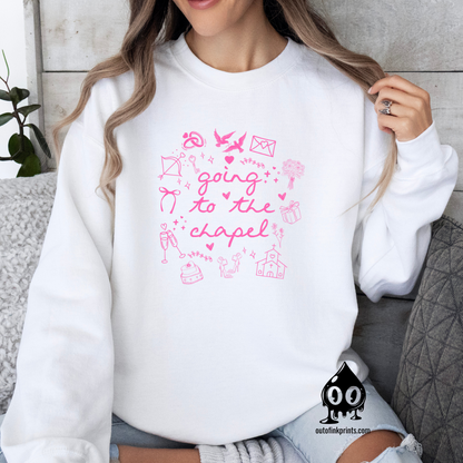 A woman sitting and wearing a white sweatshirt with pink text that reads 'Going to the Chapel' along with various symbols and text related to weddings.