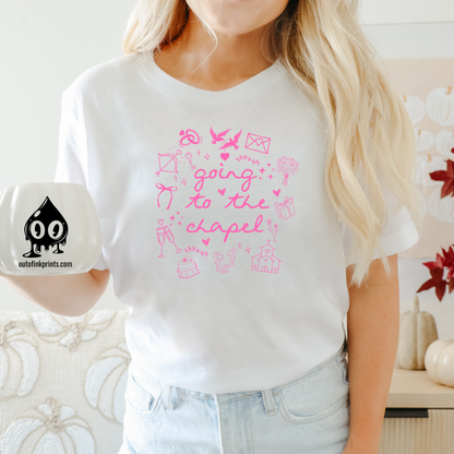 A person holding a white T-shirt with pink text and graphics related to a bride and chapel, with a coffee mug in the foreground.