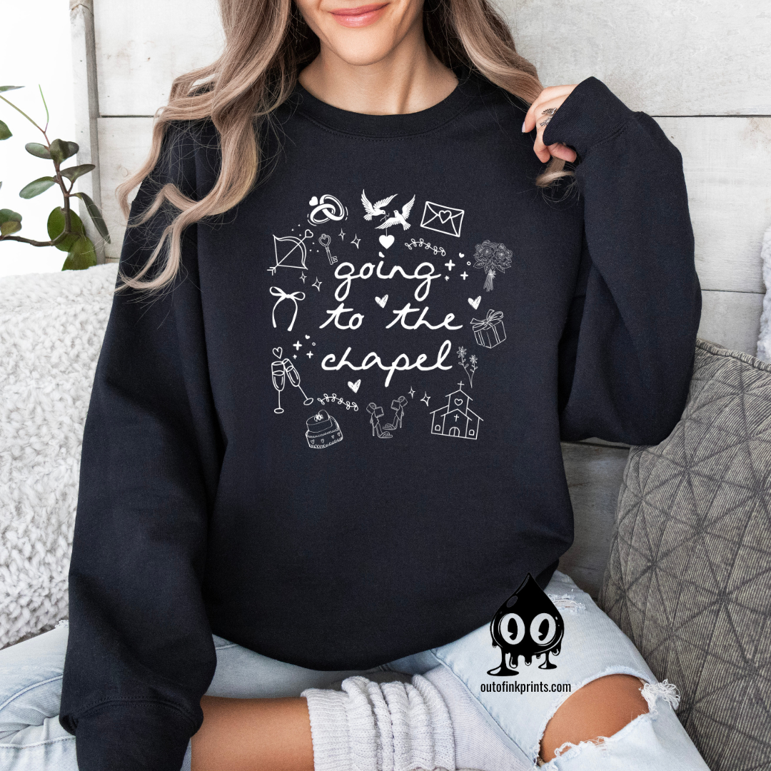 A woman sitting wearing a black sweatshirt with white text that reads 'GOING TO THE CHAPEL' and various illustrations such as a bride, groom, and wedding-related icons.