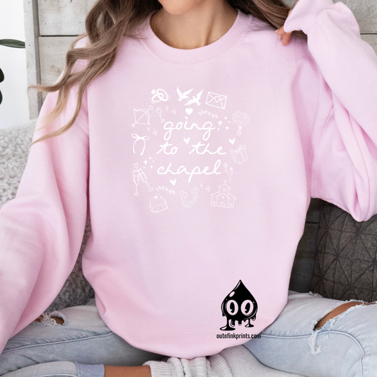 A woman sitting wearing a pink sweatshirt with white text that reads 'GOING TO THE CHAPEL' and various illustrations such as a bride, groom, and wedding-related icons.