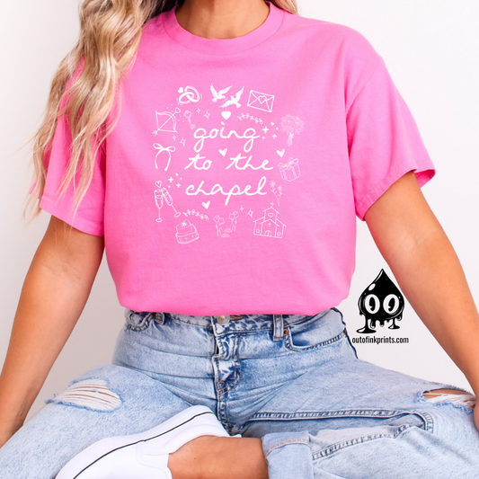 A woman sitting and wearing a pink T-shirt with white text that reads 'Going to the Chapel' along with various symbols and text related to weddings.