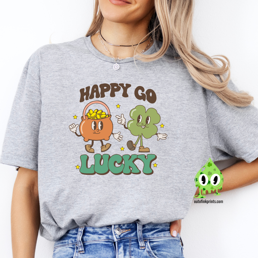 A person wearing a gray Happy Go Lucky' t-shirt with cartoonish characters and text on it, suitable for St. Patrick's Day celebrations.