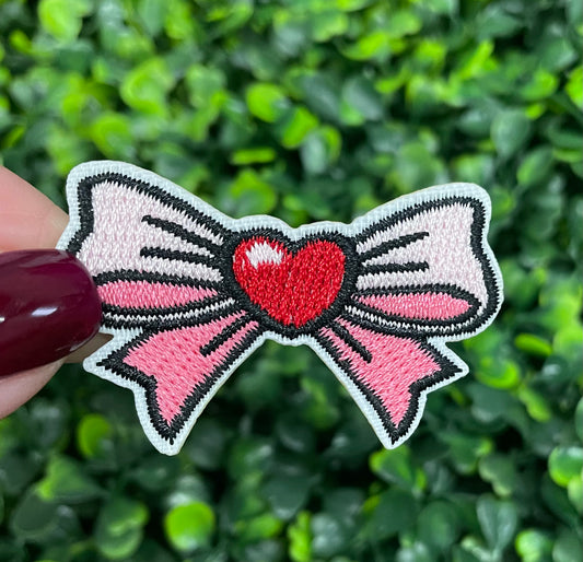 A patch featuring a pink bow with a red heart embroidered in the center, held against a green leafy background.