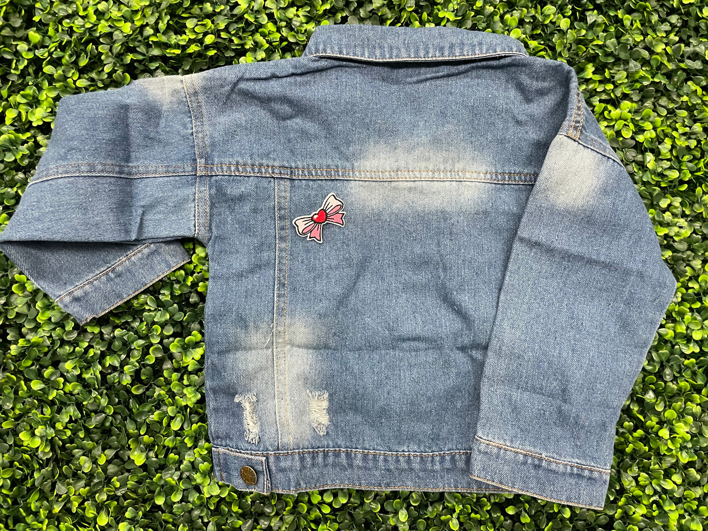 Blue jean jacket with a patch attached to the back that has a pink and white bowtie design with a heart in the center
