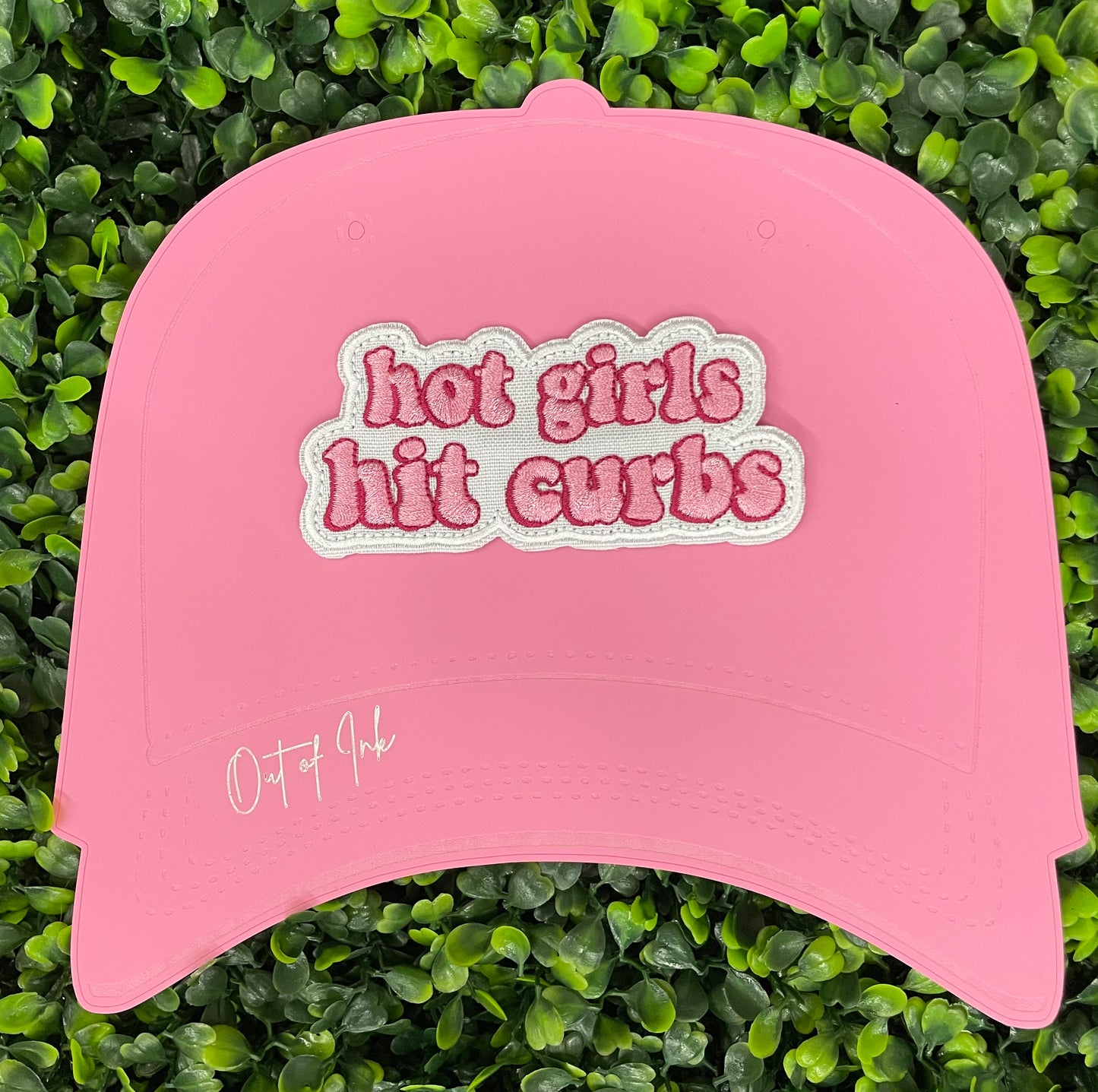 Pink hat template with an pink embroidered patch that says hot girls hit curbs with a green leafy background. 