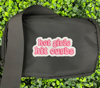 black fanny pack with an pink embroidered patch that says hot girls hit curbs with a green leafy background. 