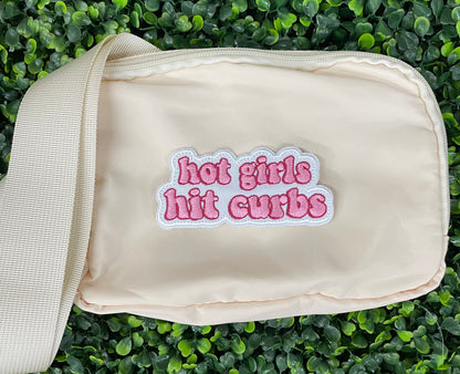 White fanny pack with an pink embroidered patch that says hot girls hit curbs with a green leafy background. 