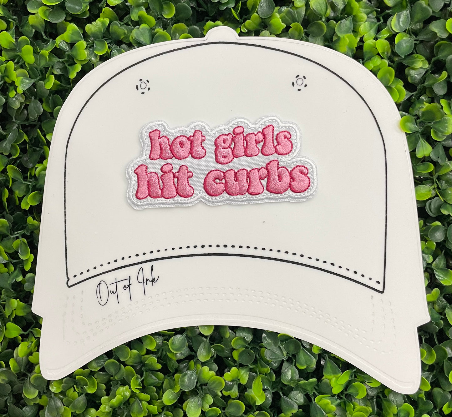 white hat template with an pink embroidered patch that says hot girls hit curbs with a green leafy background. 