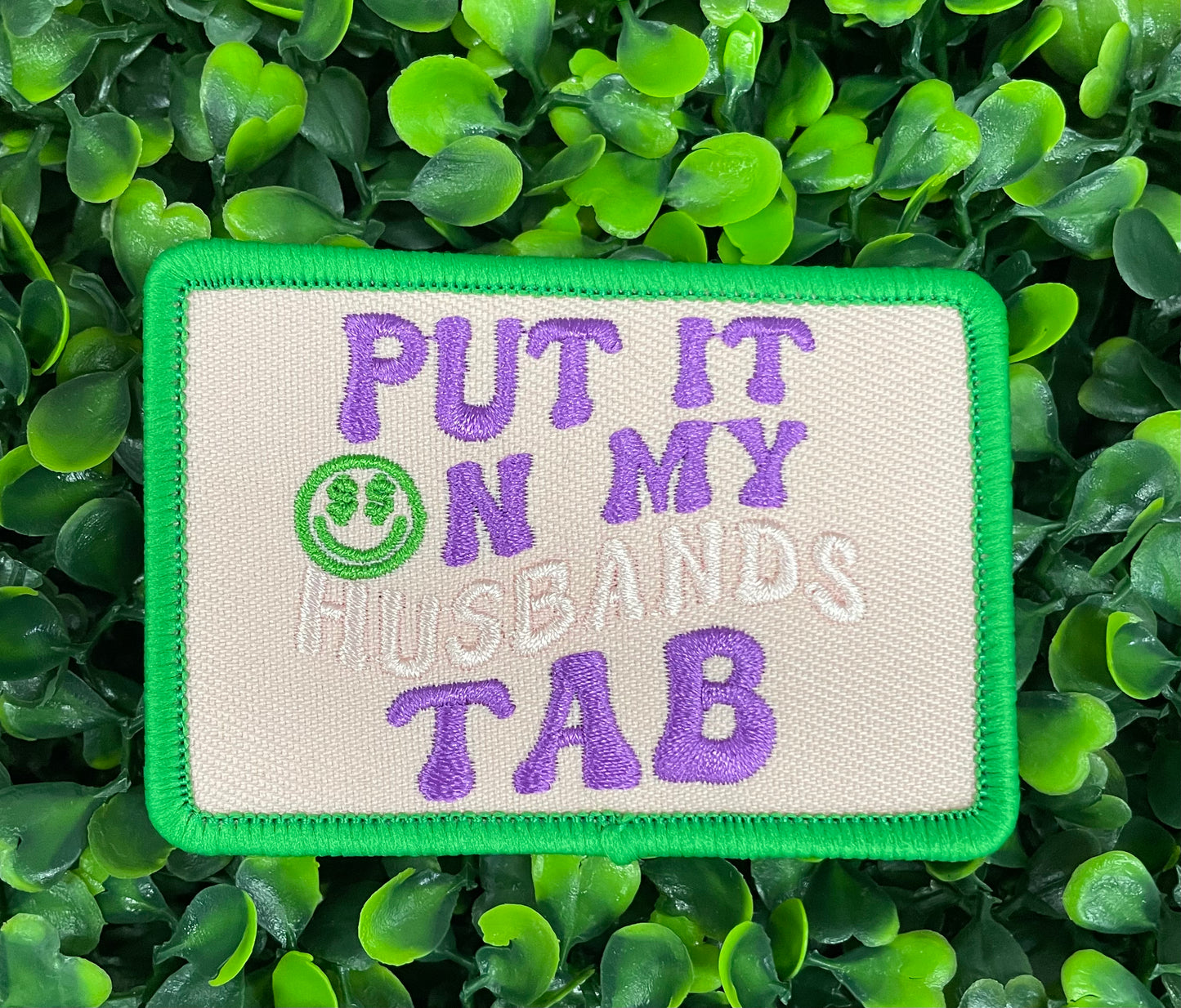 an embroidered patch with the text 'PUT IT ON MY HUSBAND'S TAB' on a green background