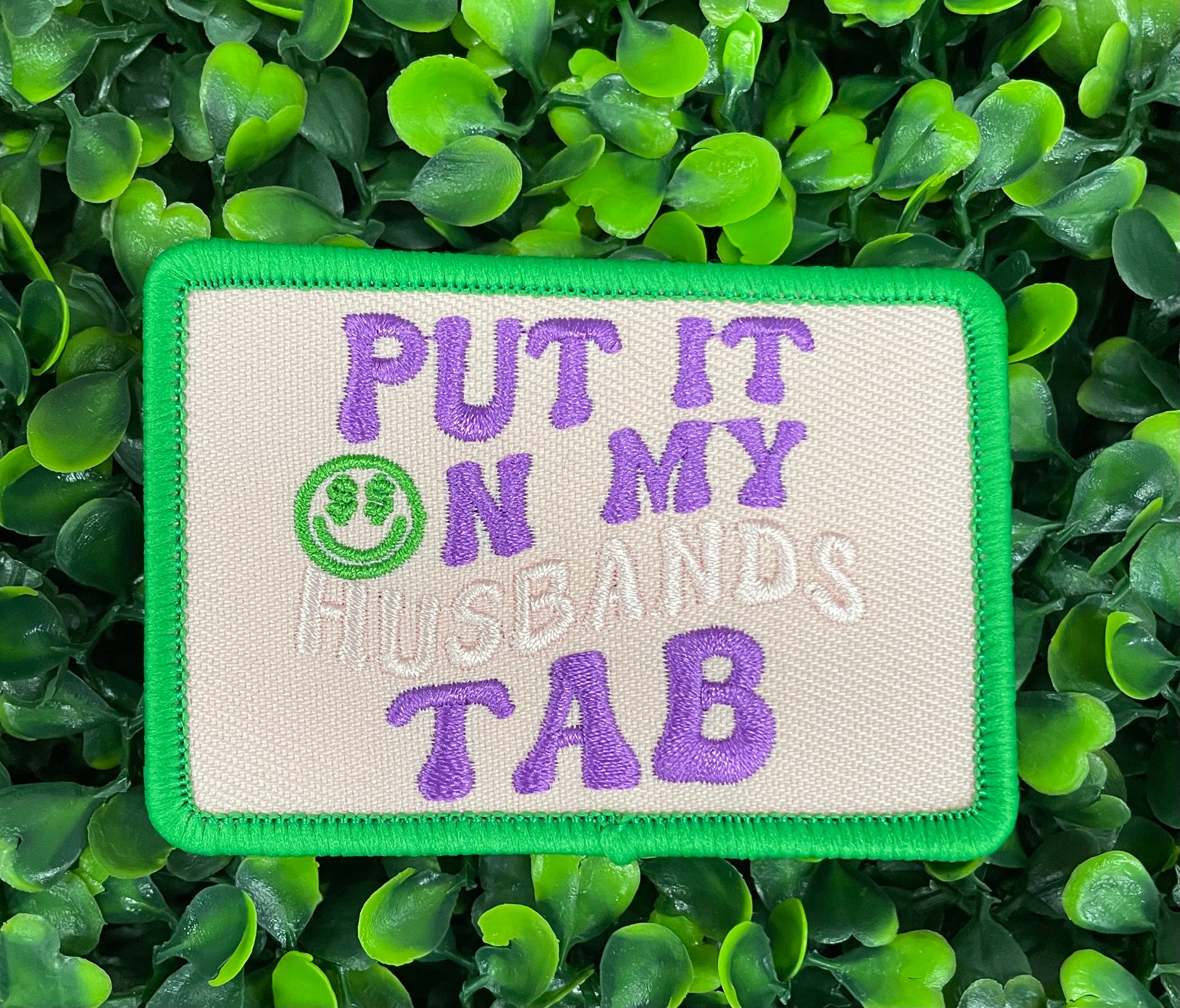 an embroidered patch with the text 'PUT IT ON MY HUSBAND'S TAB' on a green background