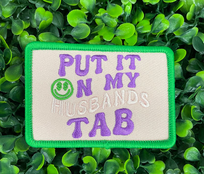 an embroidered patch with the text 'PUT IT ON MY HUSBAND'S TAB' on a green background