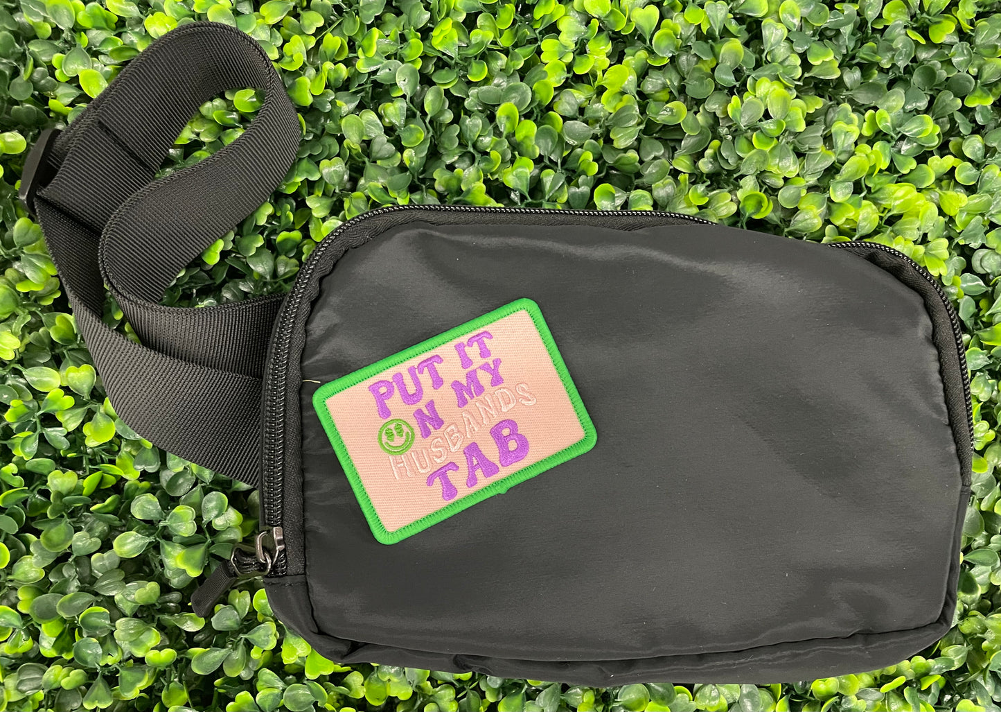  an embroidered patch with the text 'PUT IT ON MY HUSBAND'S TAB' on a green background and a black fanny pack