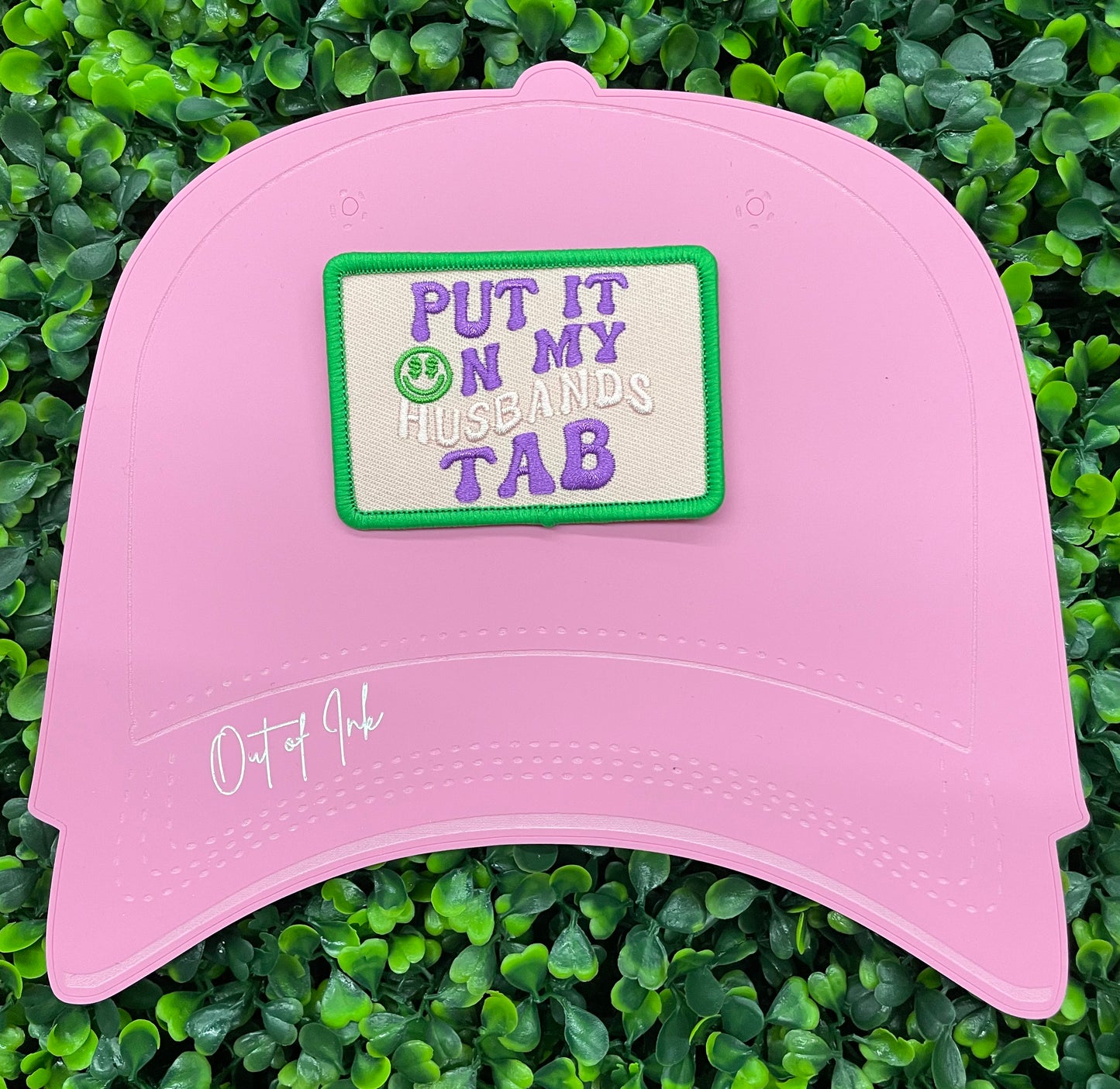 A pink hate with an embroidered patch with the text 'PUT IT ON MY HUSBAND'S TAB' on a green background 