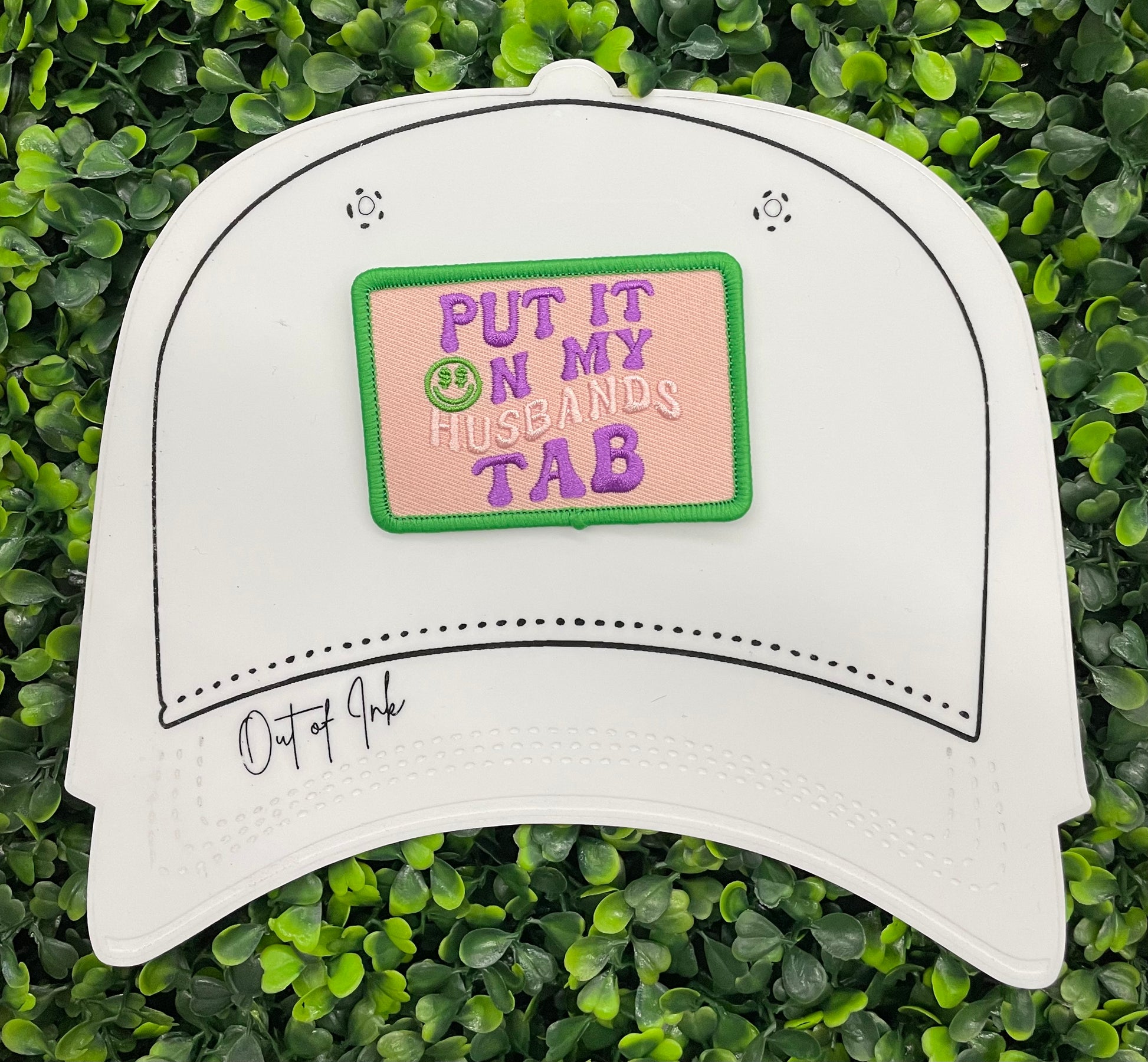 A white hat with an embroidered patch with the text 'PUT IT ON MY HUSBAND'S TAB' on a green background