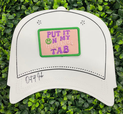 A white hat with an embroidered patch with the text 'PUT IT ON MY HUSBAND'S TAB' on a green background