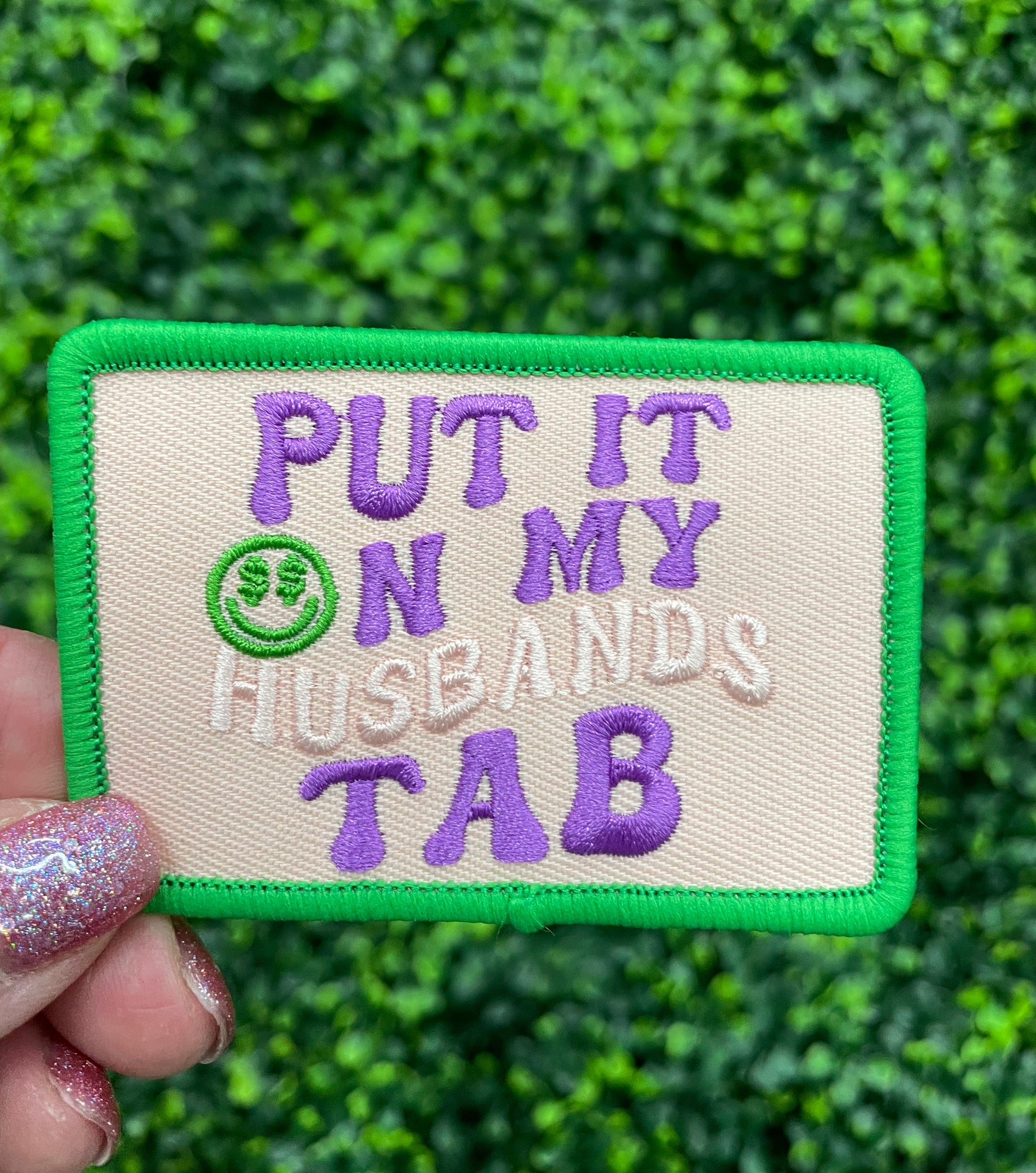 A hand holding an embroidered patch with the text 'PUT IT ON MY HUSBAND'S TAB' on a green background with a purple border.