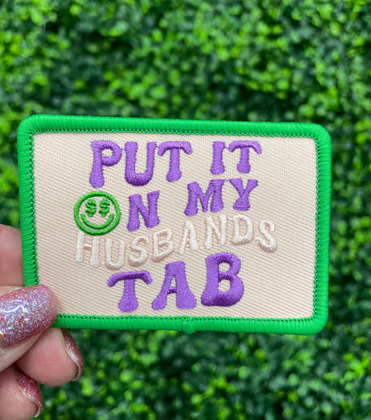 A hand holding an embroidered patch with the text 'PUT IT ON MY HUSBAND'S TAB' on a green background with a purple border.