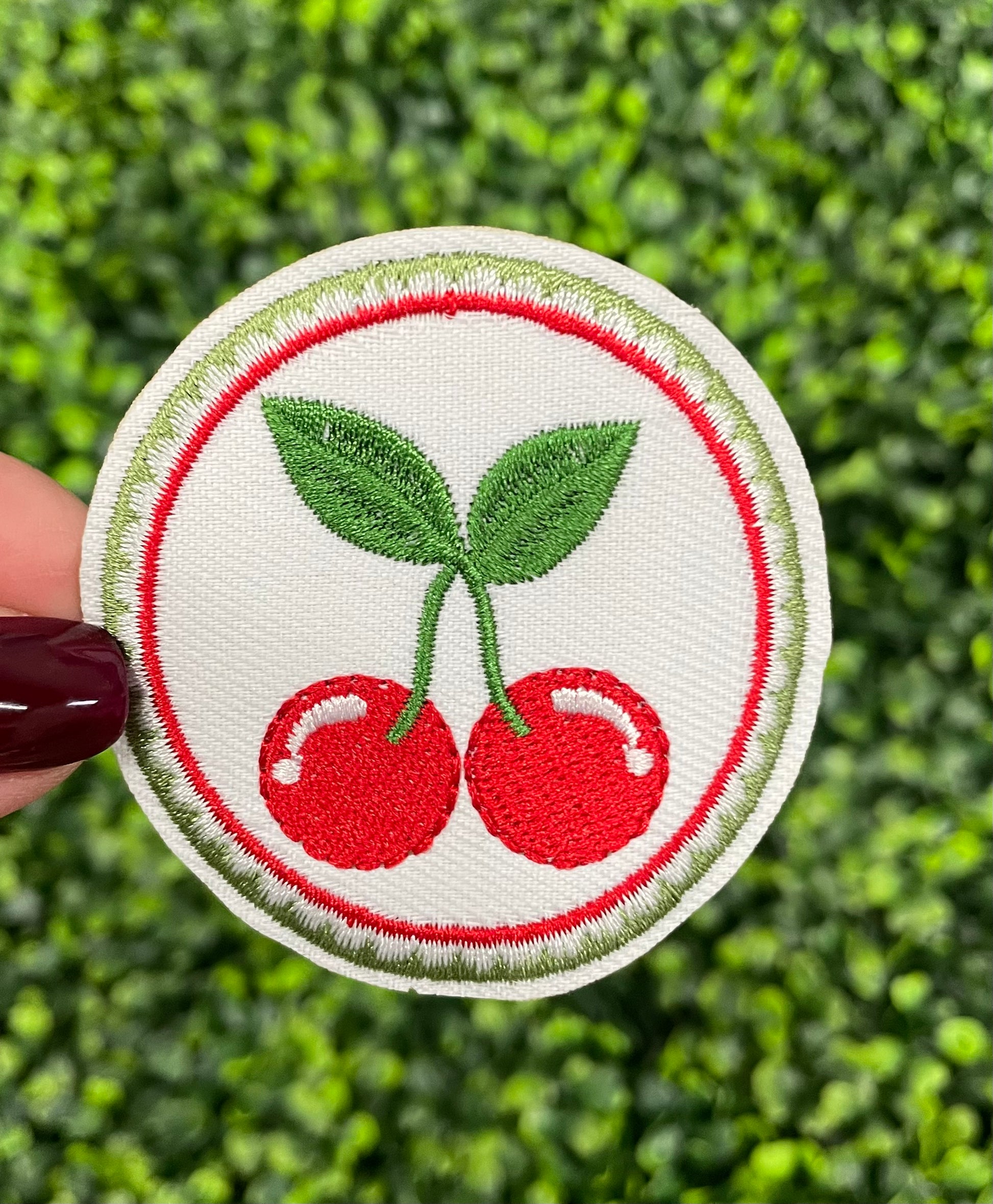 A round embroidered patch with two red cherries and green leaves on a white background, held against a green leafy backdrop.