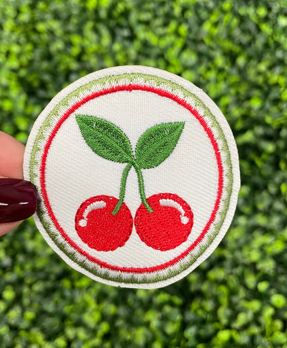 A round embroidered patch with two red cherries and green leaves on a white background, held against a green leafy backdrop.