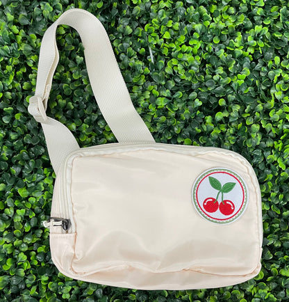 A round embroidered patch with two red cherries and green leaves on a white background, on a white fanny pack