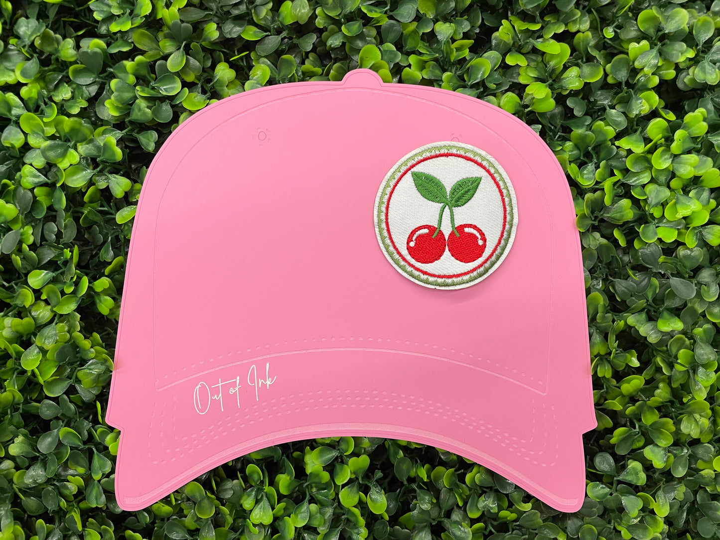 A round embroidered patch with two red cherries and green leaves on a white background, on a pink hat template