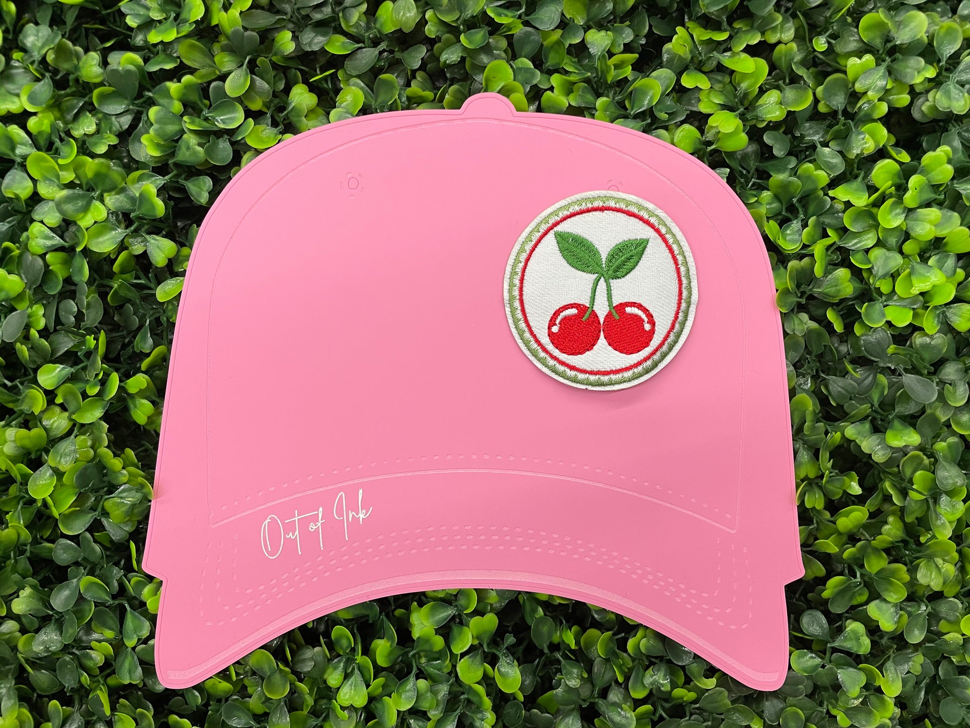 A round embroidered patch with two red cherries and green leaves on a white background, on a pink hat template