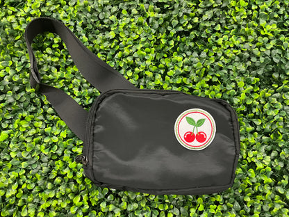 A round embroidered patch with two red cherries and green leaves on a white background, on a black fanny pack