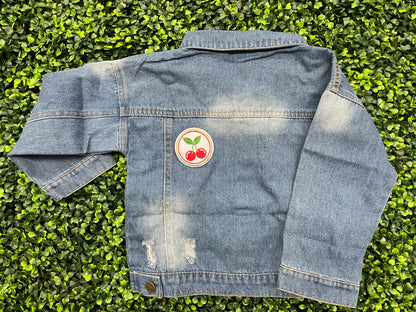 A round embroidered patch with two red cherries and green leaves on a white background, on a jean jacket