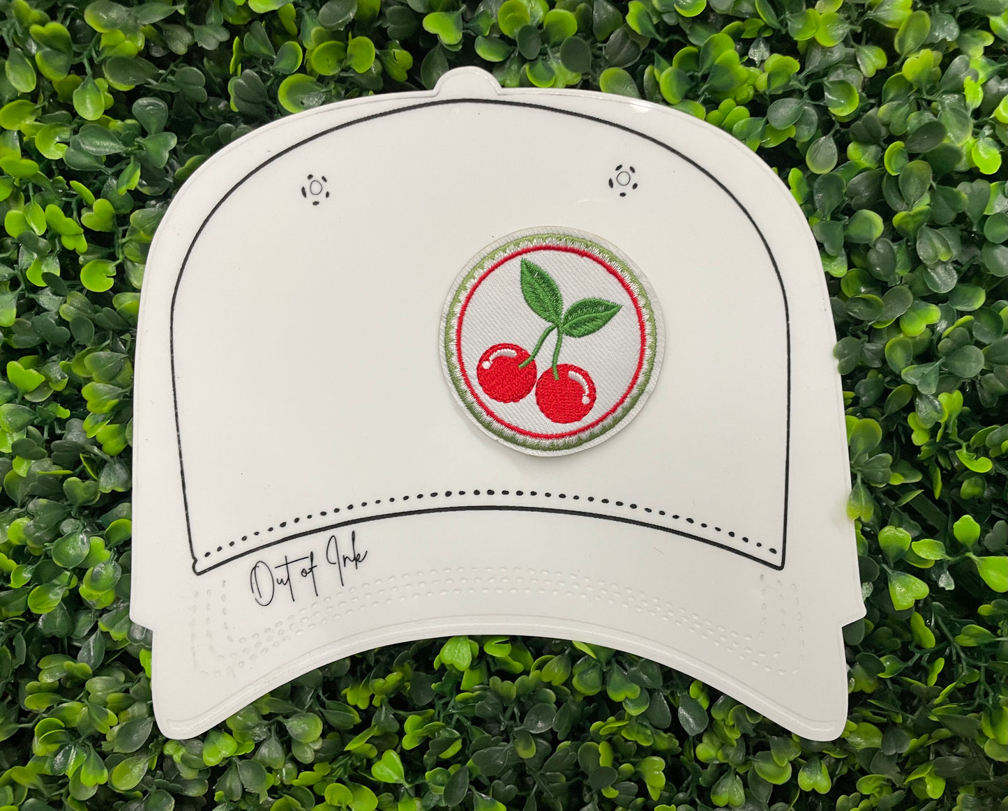 A round embroidered patch with two red cherries and green leaves on a white background, on a white hat template