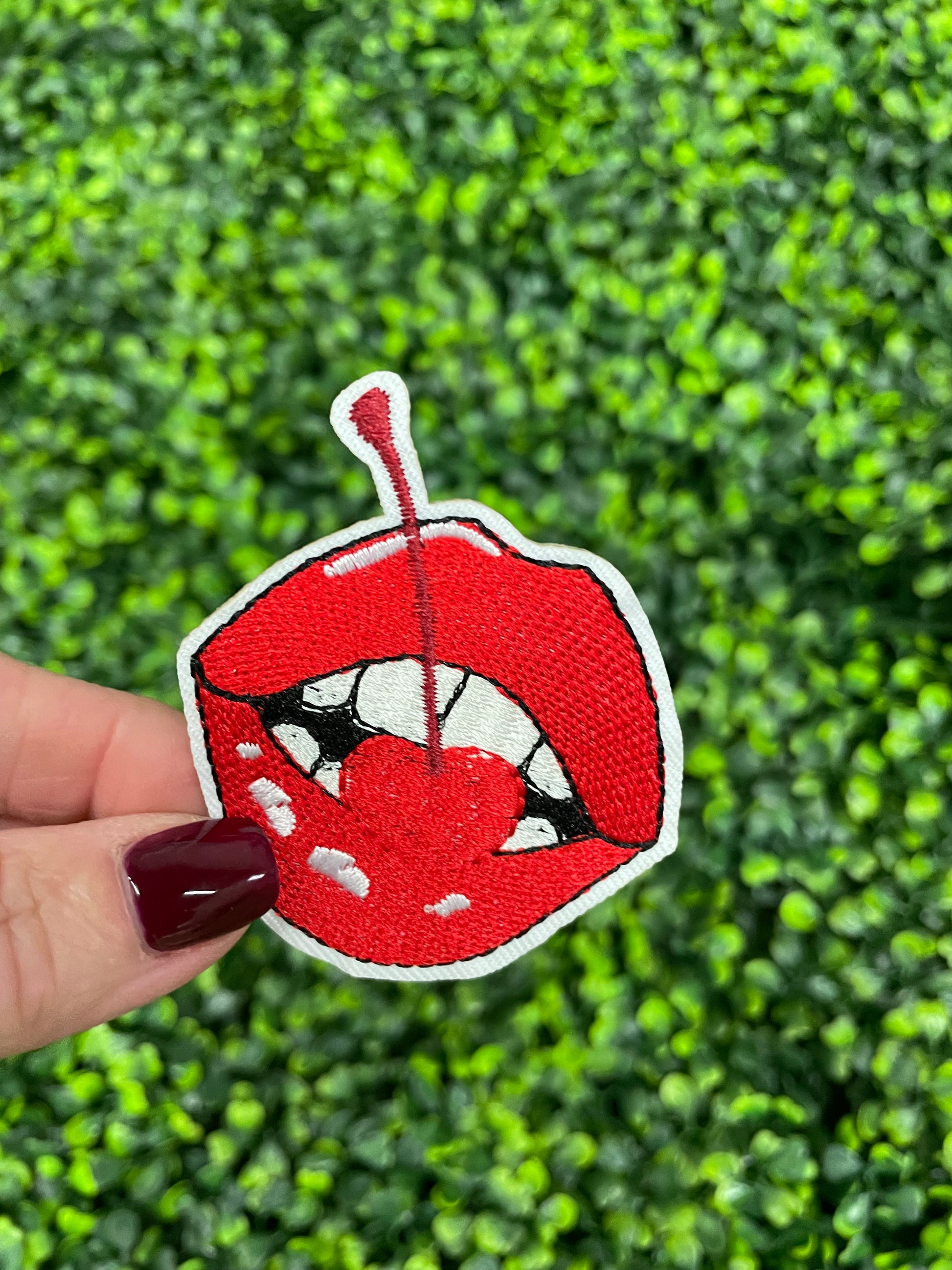 A person holding a red lips embroidered patch with a cherry, with greenery in the background.