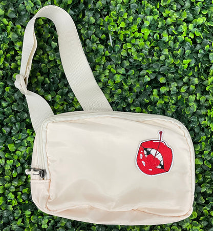 white fanny pack with a red lips embroidered patch with a cherry, with greenery in the background.