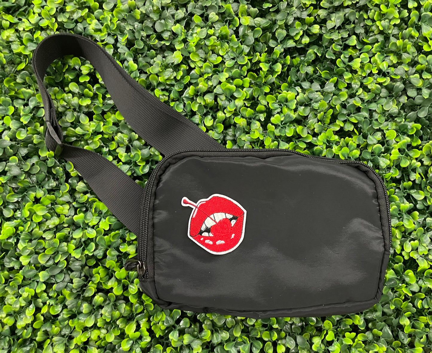 black fanny pack with a red lips embroidered patch with a cherry, with greenery in the background.