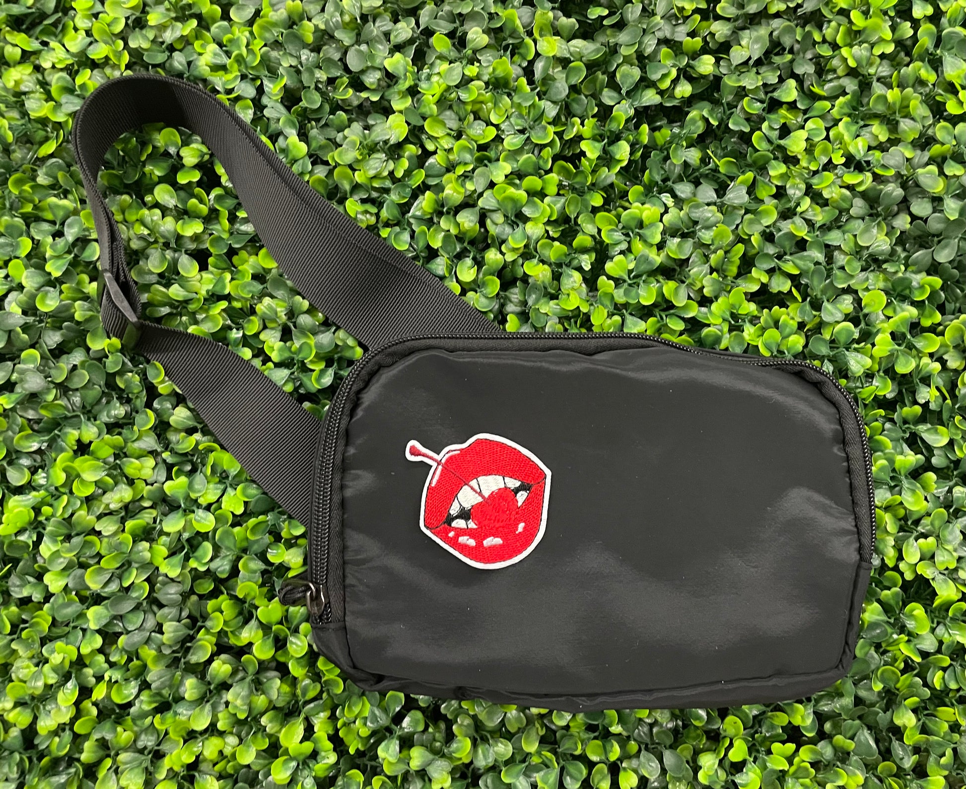 black fanny pack with a red lips embroidered patch with a cherry, with greenery in the background.